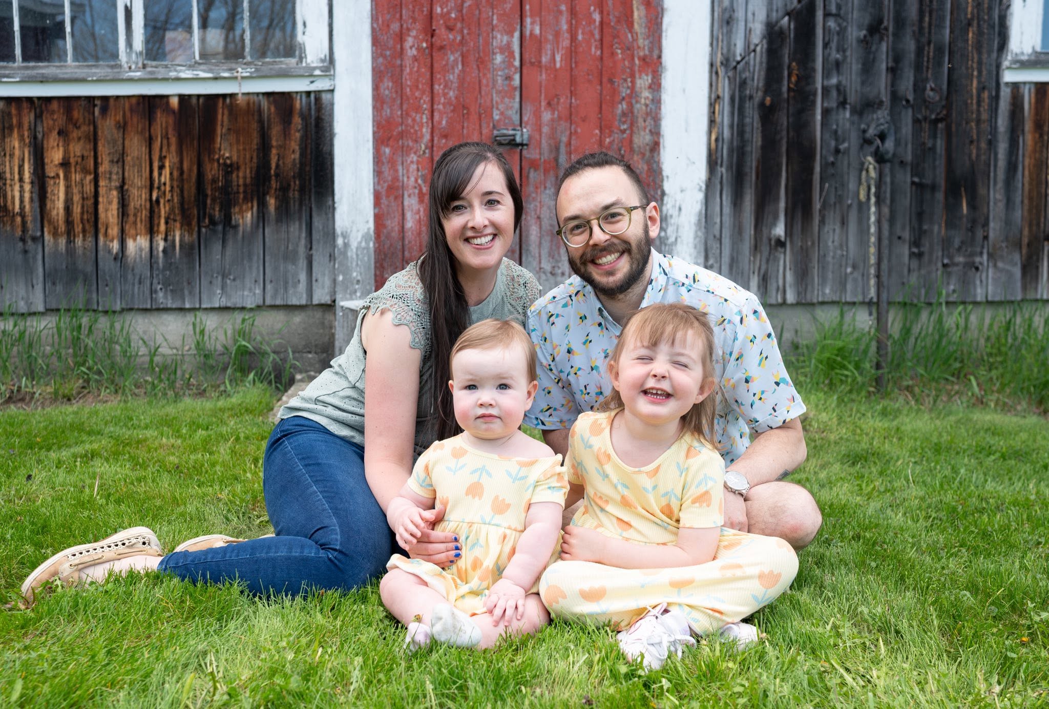 A family of four laughing together on the grass in front of a weathered red barn